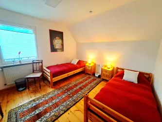 Ein einfaches Zimmer mit zwei Einzelbetten und einem Teppich. Es gibt ein Fenster, das natürliches Licht hereinlässt.