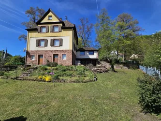 Ein großes, zweigeschossiges Haus mit einem Garten und bunten Blumen. Im Hintergrund sind Bäume und ein blauer Himmel sichtbar.
