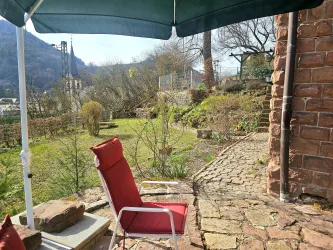 Eine sonnige Sitzecke im Garten mit einem roten Stuhl und einem Sonnenschirm. Im Hintergrund sieht man üppiges Grün und alte Steinmauern.
