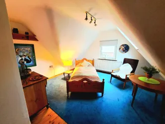 Ein gemütliches Schlafzimmer mit einem einzelnen Bett und einem Sessel. Der Raum hat einen blauen Teppich und sanfte Beleuchtung.