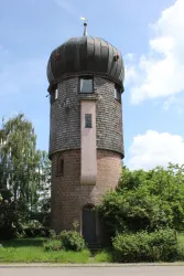 Ein historischer Wasserturm mit einer runden, kupferfarbenen Kuppel. Umgeben von Bäumen und einem klaren blauen Himmel.