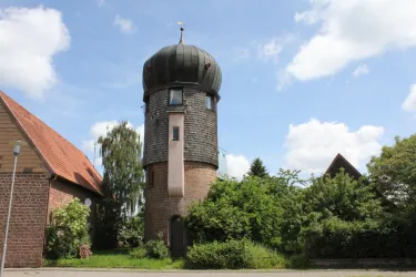 Ein historischer Wasserturm mit einer markanten Kuppel steht zwischen mehreren Gebäuden. Umgeben von Bäumen und Sträuchern strahlt die Szene eine ländliche Atmosphäre aus.