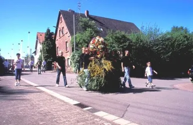 Eine festliche Parade mit Blumenkränzen auf der Straße. Menschen gehen zusammen und feiern in einer ländlichen Umgebung.