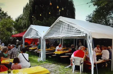 Ein gemütliches Gartenfest unter Zelten. Viele Menschen sitzen an langen Tischen und genießen die Atmosphäre.