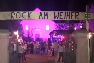 Ein Eingang zu einem Musikfestival mit einem Schild "Rock am Weiher". Im Hintergrund sind Menschen versammelt und ein hübsches Gebäude in lila Licht beleuchtet.