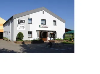 Ein weißes Gebäude mit der Aufschrift „TUS 1890“ an der Fassade. Es gibt einen kleinen Gartenbereich mit Tischen und Stühlen.