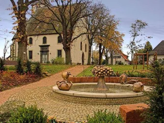 Ein schöner Platz mit einem Brunnen und einer ansprechenden Umgebung. Im Hintergrund ist ein historisches Gebäude und Bäume zu sehen.