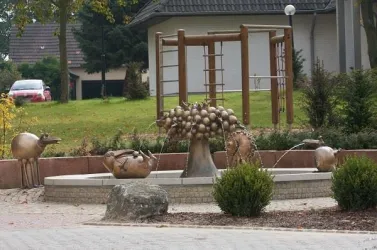 Ein kreativer Brunnen mit verschiedenen Tierfiguren aus Bronze. Im Hintergrund befindet sich ein Spielplatz und eine grüne Wiese.