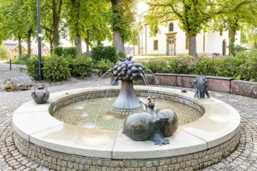 Ein kunstvoller Brunnen mit Skulpturen von Früchten und Tieren. Umgeben von Bäumen und einem ruhigen Platz.