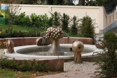 Ein moderner Brunnen mit stilisierten, bronzeartigen Skulpturen in einem Garten. Umgeben von grünen Pflanzen und Steinmauern.