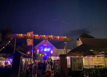 Eine lebendige Veranstaltung mit vielen Menschen unter dem Schild „Rock am Wehner“. Bunte Lichter erleuchten die Umgebung bei Dämmerung.