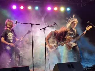 Eine Rockband spielt live auf einer Bühne mit bunten Lichtern. Die Musiker sind in Bewegung und von Nebel umgeben.