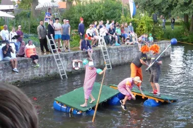 Eine Gruppe von Clowns steht auf einer schwimmenden Plattform im Wasser. Zuschauer sitzen am Ufer und beobachten das Geschehen.