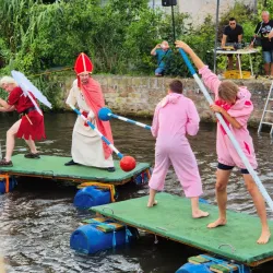 Eine Gruppe von Personen in bunten Kostümen kämpft auf schwimmenden Plattformen im Wasser. Im Hintergrund sind Zuschauer und ein festliches Ambiente zu sehen.