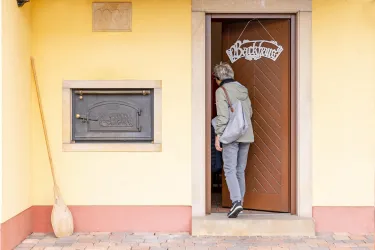 Eine Person tritt durch die Tür eines Hauses mit einer Schiefertafel, auf der "Backhaus" steht. Neben der Tür befindet sich ein traditioneller Ofen und ein Holzlöffel.