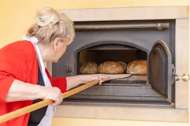 Eine Frau zieht frisches Brot aus einem Holzofen. Sie hält ein großes Holzbrett in der Hand.