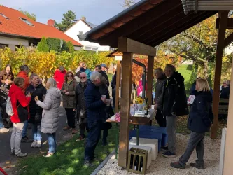 Eine große Gruppe von Menschen steht in einem Gartenbereich und unterhält sich. Im Hintergrund sind bunte Bäume und ein Holzunterstand zu sehen.