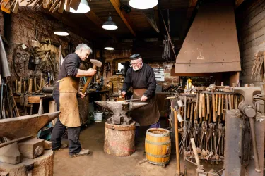 Zwei Schmiede arbeiten in einer traditionellen Werkstatt. Der eine bearbeitet ein Stück Metall am Amboss, während der andere assistiert.