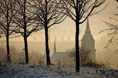 Eine winterliche Landschaft mit schneebedecktem Boden und silhouettierten Bäumen. Im Hintergrund ist eine Kirche mit einem spitzen Turm zu sehen, umhüllt von Nebel.