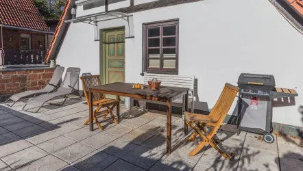 Eine gemütliche Terrasse mit einem Holztisch und Stühlen. Im Hintergrund steht ein Grill und es gibt Liegen zum Entspannen.