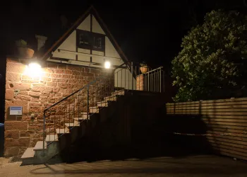 Ein charmantes, beleuchtetes Haus mit einer Steintreppe bei Nacht. Bäume und Pflanzen umgeben den Eingang und verleihen der Szene eine gemütliche Atmosphäre.