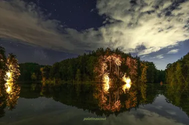 Eine ruhige Landschaft mit einem See, umgeben von Bäumen und beleuchtet von sanften Lichtern. Der Nachthimmel ist mit Wolken und Sternen geschmückt.