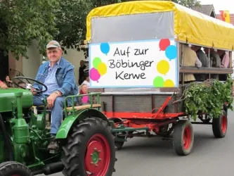 Ein Mann sitzt auf einem grünen Traktor, der einen Wagen mit einer bunten Banner fährt. Das Banner wirbt für die Böbinger Kerwe mit festlichen Farben.