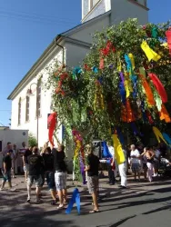 Eine festliche Szenerie mit einer großen, geschmückten Baumdekoration und bunten Bändern. Menschen feiern auf der Straße in der Nähe eines Gebäudes.