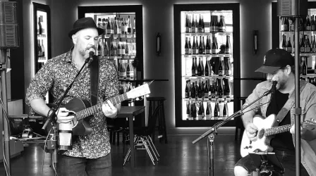 Zwei Musiker spielen live in einem stilvollen Raum. Im Hintergrund sind Regale mit verschiedenen Getränken zu sehen.