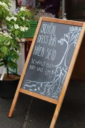 Ein Holztäfelchen mit der Aufschrift „Schön, dass ihr hier seid! Schaut euch bei uns um!“. Es ist umgeben von Pflanzen und vermittelt eine einladende Atmosphäre.