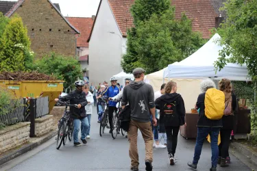 Eine belebte Straße mit verschiedenen Menschen, die Fahrräder benutzen und sich um einen Marktstand versammeln. Im Hintergrund sind Häuser und grüne Bäume zu sehen.