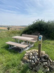 Ein Picknickplatz mit einer Holztischgruppe auf einer Wiese. Im Hintergrund sind sanfte Hügel und ein klarer Himmel zu sehen.