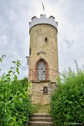Ein historischer Turm aus Stein mit einem rot-weißen Dach. Umgeben von üppigem Grün und einem Treppenaufgang, der zum Eingang führt.