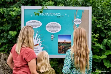 Drei Kinder stehen vor einem informativen Schild über den „Furnasenweg“. Das Schild enthält Texte und Bilder, um den Weg und die Umgebung zu präsentieren.