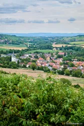 Eine malerische Landschaft mit einem kleinen Dorf und einer Kirche im Vordergrund. Sanfte Hügel und grüne Felder umgeben die Szenerie.