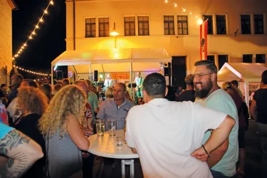 Eine lebhafte Veranstaltung bei Nacht mit vielen Menschen, die miteinander sprechen und Spaß haben. Im Hintergrund spielt eine Band, und die Atmosphäre ist festlich.