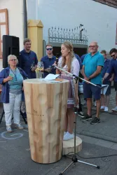 Eine junge Frau spricht an einem Rednerpult vor einer Gruppe von Menschen. Im Hintergrund sind ein paar Personen mit Getränken und Musikinstrumenten zu sehen.