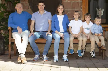 Eine Familie sitzt gemeinsam auf einer Bank im Freien. Sie lächeln in die Kamera und tragen bequeme Kleidung.
