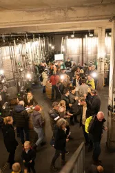 Eine belebte Weinkellerei mit vielen Menschen, die sich unterhalten und Wein probieren. Im Hintergrund sind Weinfässer und große Lagereinrichtungen zu sehen.