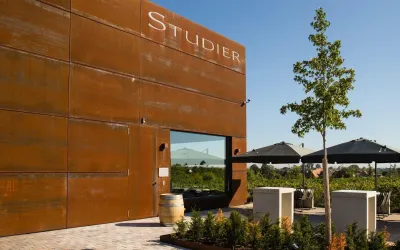 Ein modernes Gebäude mit der Aufschrift "Studier" steht im Vordergrund. Im Hintergrund sind Weinreben und Sonnenschirme zu sehen.
