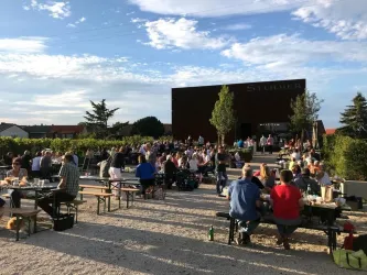 Eine lebhafte Veranstaltung im Freien mit vielen Menschen, die an Tischen sitzen und essen. Im Hintergrund sieht man ein modernes Gebäude und Weinreben.