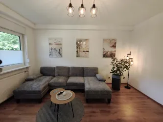 Ein modernes Wohnzimmer mit einem grauen Sofa und einem runden Tisch. An den Wänden hängen Bilder, und eine Pflanze steht in der Ecke.
