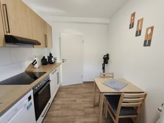 Eine moderne Küche mit Holzschränken und einer hellen Arbeitsfläche. An einem kleinen Tisch stehen drei Stühle und an der Wand hängen einige Bilder.