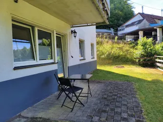 Eine Terrasse mit einem schwarzen Tisch und zwei Stühlen vor einem Wohnhaus. Im Hintergrund ist eine grüne Wiese und einige Bäume sichtbar.