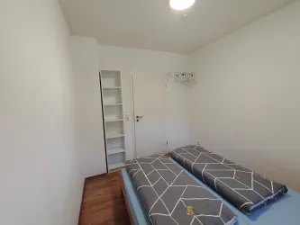Ein einfaches Zimmer mit zwei Betten und einem Regalschrank. Die Wände sind schlicht und die Böden aus Holz.
