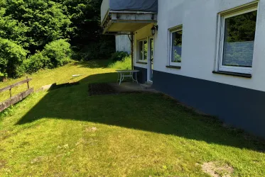 Ein Haus mit einem grünen Garten und einer Terrasse. Im Hintergrund sind Bäume und eine ruhige Landschaft zu sehen.