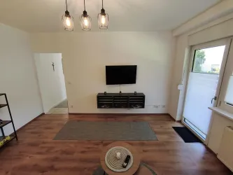 Ein modernes Wohnzimmer mit einem Fernseher an der Wand und einem gemütlichen Teppich. Große Fenster sorgen für viel Tageslicht und einen Blick nach draußen.