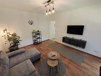 Ein modernes Wohnzimmer mit einem grauen Sofa und einem Fernseher an der Wand. Es gibt einen kleinen Tisch und eine Pflanze in der Ecke.