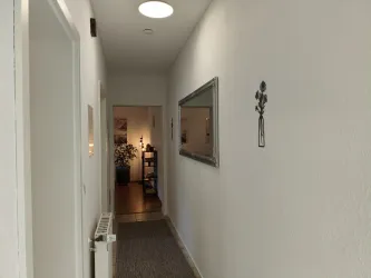Ein hell erleuchteter Flur mit weißen Wänden und einem Teppichboden. Am Ende des Flurs sind ein Spiegel und eine Pflanze sichtbar.