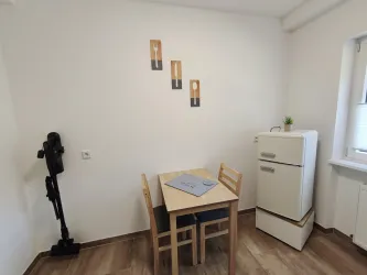 Ein kleiner, heller Raum mit einem Tisch und Stühlen. An der Wand hängen Bilder, und ein Kühlschrank steht in der Ecke.